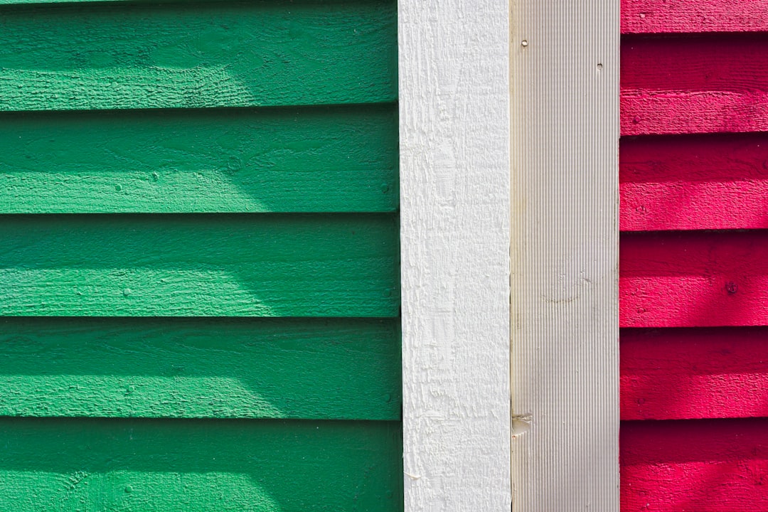 Close Up of Green, White, and Red Painted Wood Siding - A close-up image of a wooden wall with three sections of painted siding. The leftmost section is painted a vibrant green, the center section is white, and the rightmost section is bright red. The sun is shining on the wall, creating shadows.